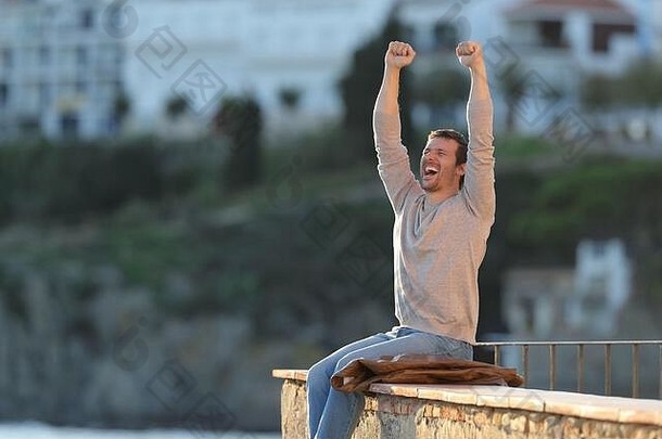 一名兴奋的男子坐在海滨小镇的阳台上举起双臂庆祝成功