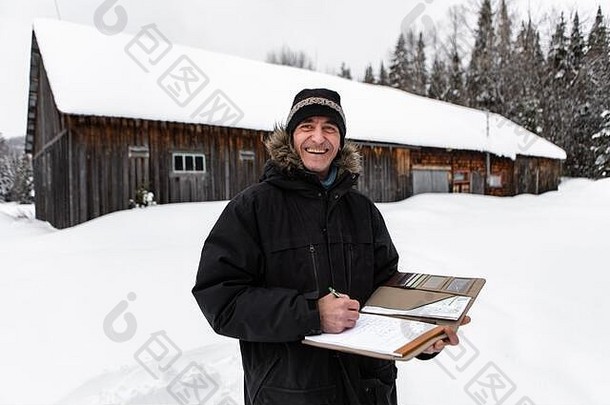 中年家居督察在巡查时做笔记。身着冬衣、面带微笑的男子站在户外雪地里，木构农舍背景