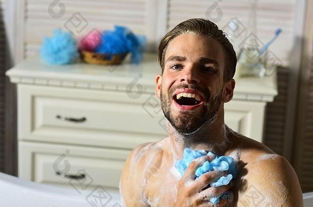留着胡子、脸上带着快乐的男人正在洗澡。男子汉光着身子坐在浴缸里用海绵洗衣服，大笑不止。健康和放松的概念。浴室里的家伙，背景是化妆品。