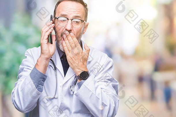 中年老年白发医生男子在孤立的背景下用智能手机说话捂住嘴，因错误而感到羞愧，表情激动