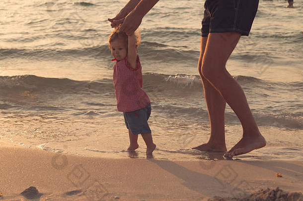 父亲帮助宝宝迈出第一步，教宝宝走路的概念，在海滩上拍摄户外照片，在海滩上度过一天，健康的家庭生活方式。