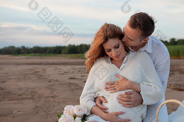 野餐、<strong>爱情</strong>、温柔。这对夫妇坐在一块格子布上，上面有枕头、鲜花和水果。一名男子从背后拥抱一名孕妇。