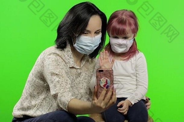 母亲、女儿戴着口罩，手持手机在视频通话。C2019冠状病毒疾病的社会疏远保护概念可爱的小孩喜欢使用智能手机。社会化媒体。色度键