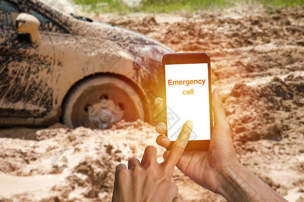 一名男子用智能手机拨打紧急求助电话，汽车轮胎陷在泥地里。车轮在泥土里跑不出来。