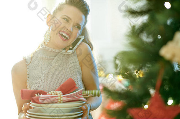微笑着的时髦家庭主妇在圣诞树旁用手机端盘子