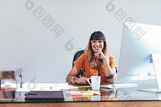 微笑的创意艺术家坐在办公室的办公桌前，在数码写字板上画草图。坐在办公室里创作自己的设计作品的插图画家