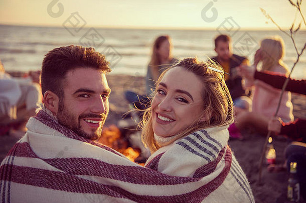 日落时分，一对年轻夫妇和朋友们在海滩上的篝火旁畅饮啤酒