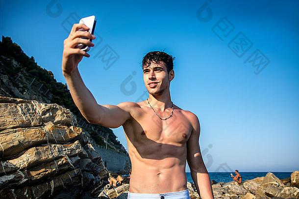 一边视图赤膊上阵年轻的男人。采取自拍照片海滩坐着大岩石热太阳