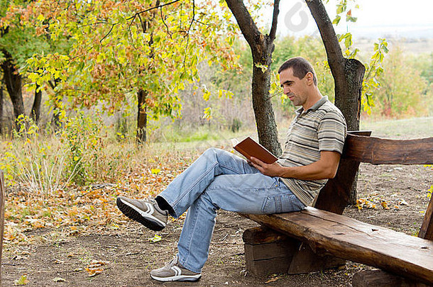 在秋天的林地里，一个男人坐在一张简陋的木凳上放松地看书