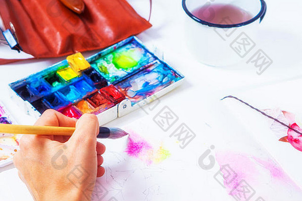 一位特写艺术家在<strong>画册</strong>中用木笔和水彩画出粉红色的花，桌上放着一个画盘，一个带画笔的皮箱