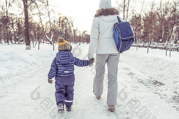 妈妈牵着儿子的手沿街走着，为他送行。后视图。冬天在城市的雪堆背景下。活动结束后回家。