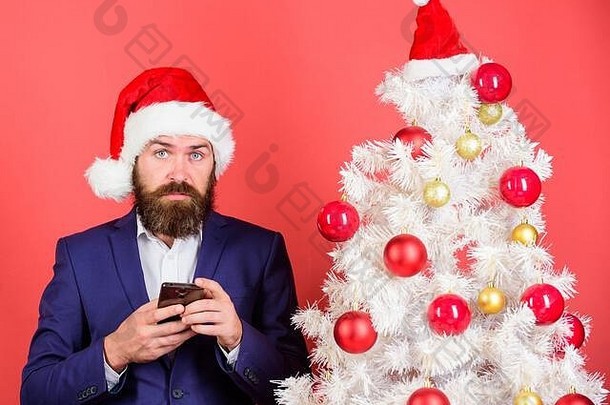 读圣诞祝福语。留着大胡子的时髦男士穿着正式西装，戴着圣诞老人的帽子。发送圣诞问候手机短信。简短的圣诞祝福。经理在网上祝贺同事。