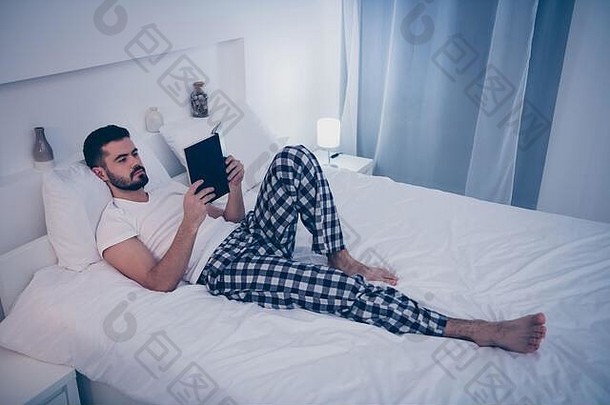 他那英俊迷人的黑发男人的肖像躺在白色的床上休息，晚上在家里酒店房间的公寓里读着有趣的书