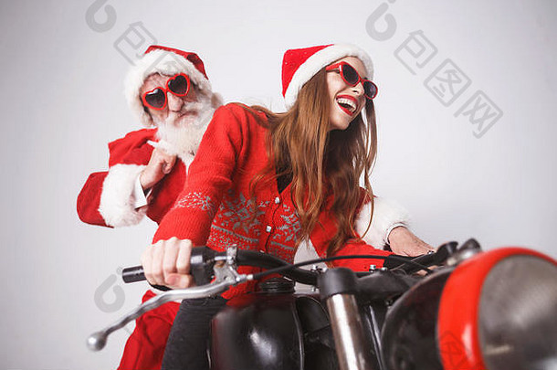 快乐的年轻圣诞老人夫人戴着圣诞帽、红色毛衣和太阳镜，一边笑着，一边骑着圣诞老人的摩托车上高速，新年、圣诞节、节假日、纪念品、礼品、<strong>购</strong>物、折扣、商店、雪人圣诞老人、化妆、发型、<strong>狂欢</strong>节。