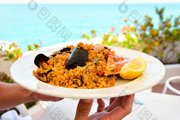 特写镜头：一个年轻人端着一盘典型的西班牙海鲜饭，带着海鲜，来到一张午餐桌旁，背景是大海