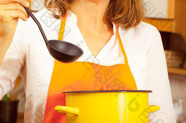 家庭主妇做汤餐晚餐。拿着勺子、勺子和锅的中年妇女。穿着橙色围裙准备食物的女管家。