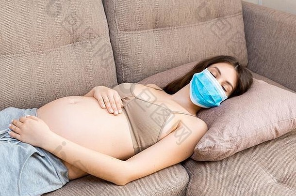 孕妇戴着医用防护面罩，以保护自己和未来的婴儿免受冠状病毒的侵害。妈妈正在家里的沙发上睡觉。全球的