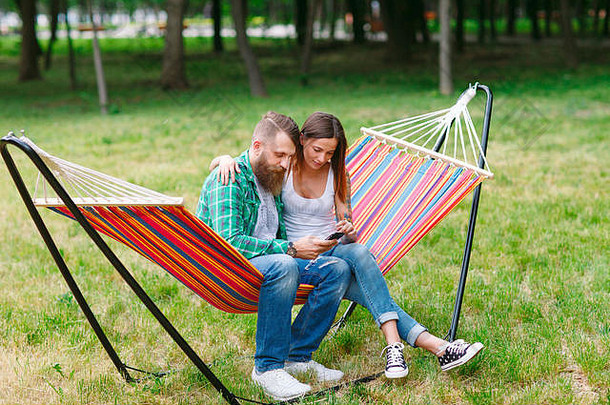 一对年轻夫妇拿着手机坐在吊床上。
