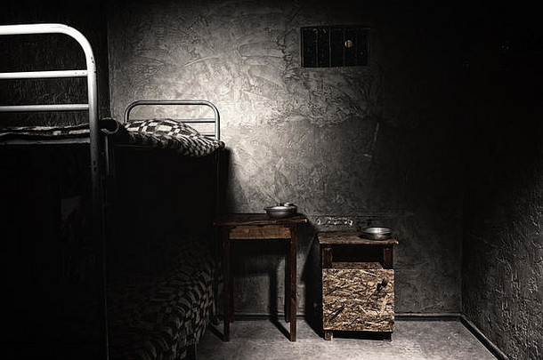 一间黑暗的空牢房，有双层床和床头柜，还有铝制盘子