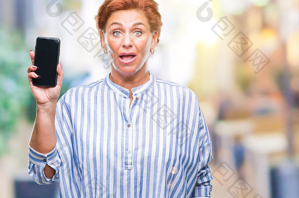 一位资深白人红发女子在孤立的背景下展示智能手机屏幕，吓得面目全非，既害怕又兴奋