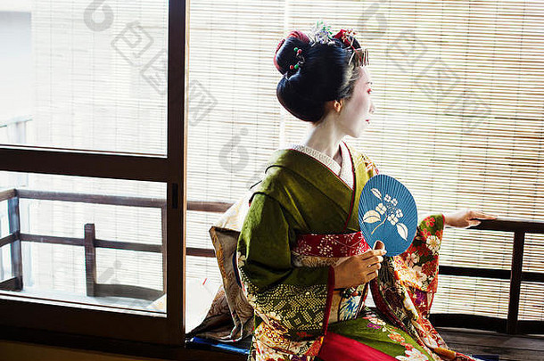 一位身着传统艺妓风格的女子，穿着和服和欧比服，拿着扇子坐在窗前。