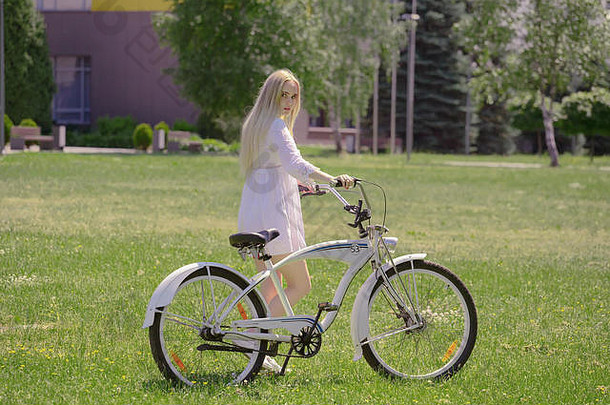 漂亮的金发碧眼的女孩白色衣服自行车绿色阳光明媚的草地走回来相机