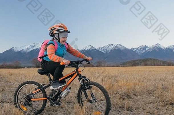 一个白人孩子在麦田里骑自行车。在美丽的雪山背景下骑着黑橙色自行车的小女孩。骑自行车的运动骑乘