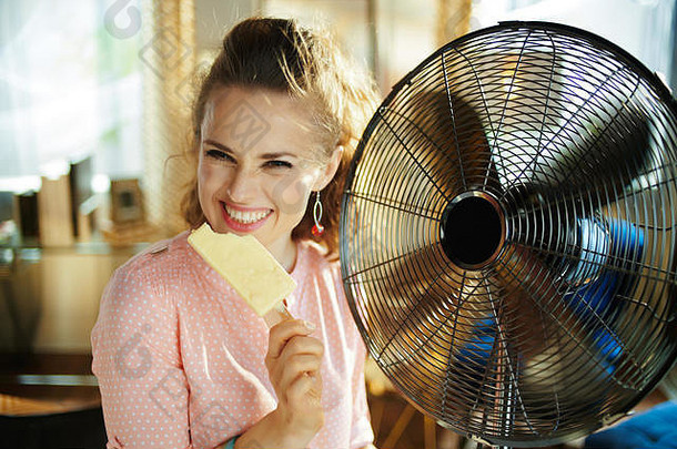 在阳光明媚、炎热的夏日，在现代化的房子里，快乐时尚的女士拿着冰激凌，用电动金属风扇降温。