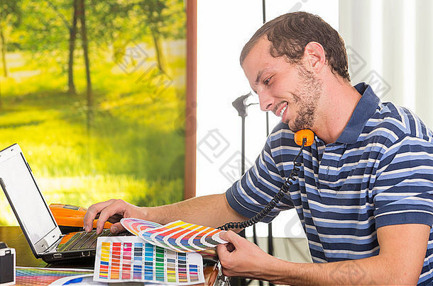 一名身穿蓝白条纹t恤的男子坐在办公桌旁用手机看调色板和彩色地图