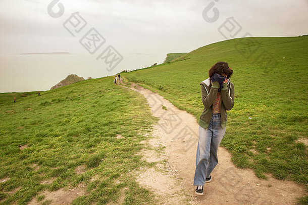 英国多塞特，一个女孩在一个刮风的日子爬上小山，<strong>保护自己</strong>的脸不受风吹
