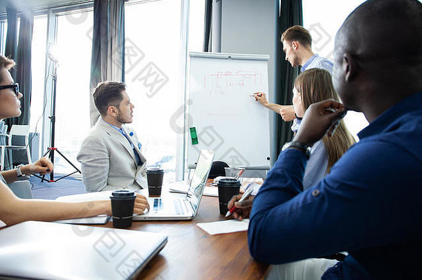 讨论公司进步自信年轻的男人。站白板指出图的同事们坐着桌子上