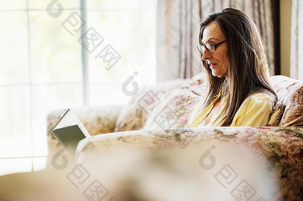 棕色长发的老妇人坐在沙发上看书，戴着阅读眼镜。