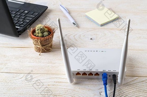 白色的Wi-Fi无线路由器靠近笔记本电脑，放在白色的木桌上。在家里或办公室的桌子上插入网线的Wlan路由器。后视图