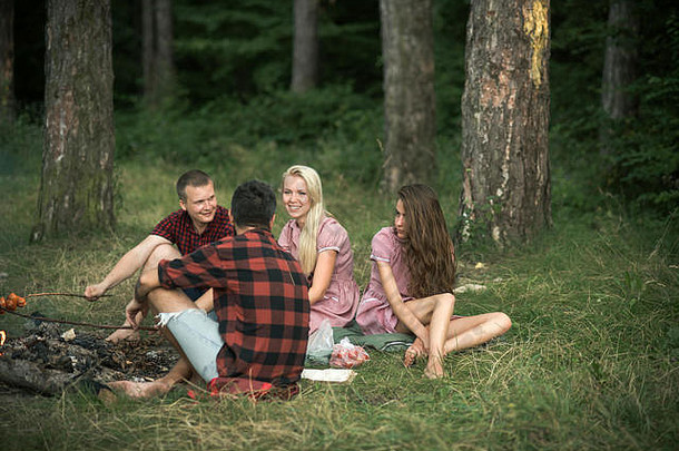 身着复古长裙的金发女孩微笑着坐在篝火旁的朋友中间。年轻人在森林野餐，友谊和休闲的概念