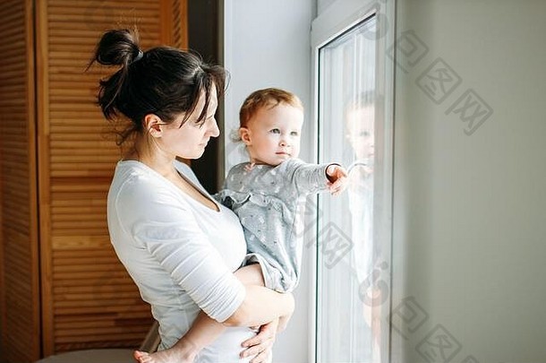 家里窗户附近，一位年轻的妈妈手上抱着一个小女孩