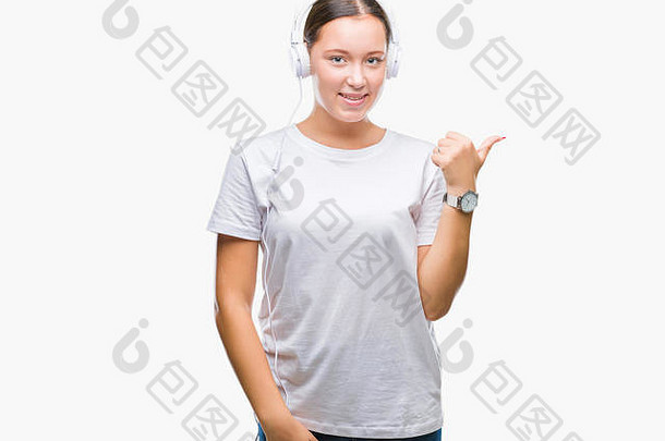 年轻的白人妇女戴着耳机在孤立的背景下听音乐，面带笑容，面带笑容，并用手指着一边