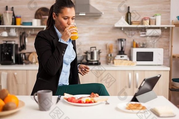 女商人在早餐时喝新鲜果汁，然后在工作前观看平板电脑上的视频。女商人上班前在网上阅读最新新闻，在厨房里使用现代技术，同时吃健康的一餐