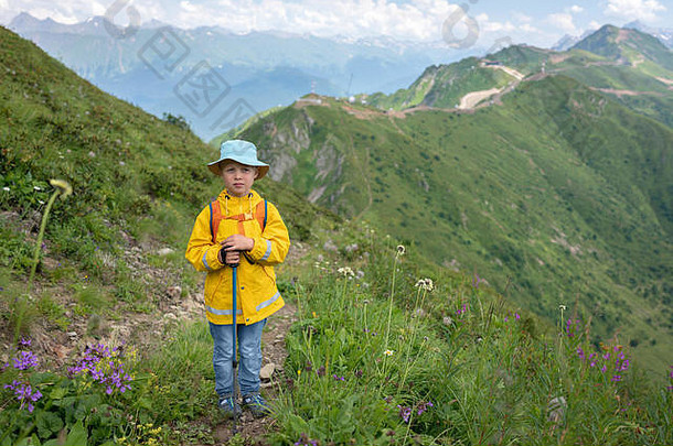一个穿着黄色雨衣和巴拿马皮鞋、手持<strong>登山杖</strong>的男孩站在通往山顶的小路上。高加索，俄罗斯