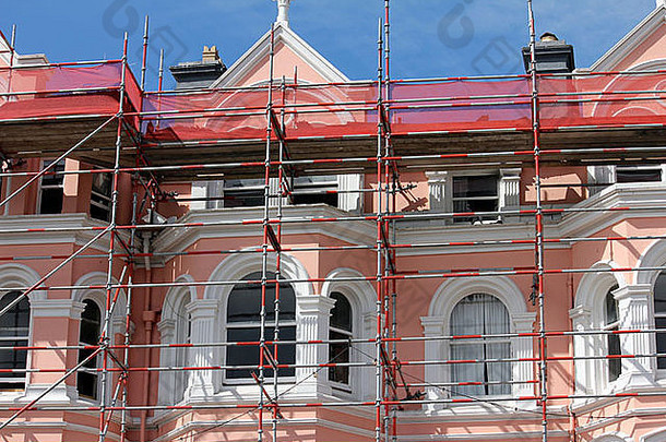 红色的金属脚手架映衬着一座粉色和白色的维多利亚式房屋，映衬着蓝天