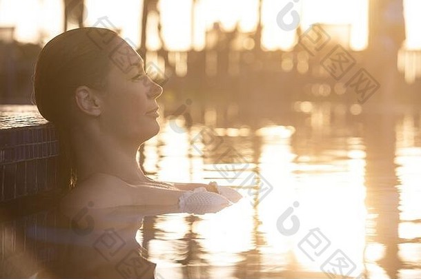 日落时在泳池边休息的女人。强烈的暖光。从侧面可以看到美丽的面部特征。灯光在水中的反射。夏季度假