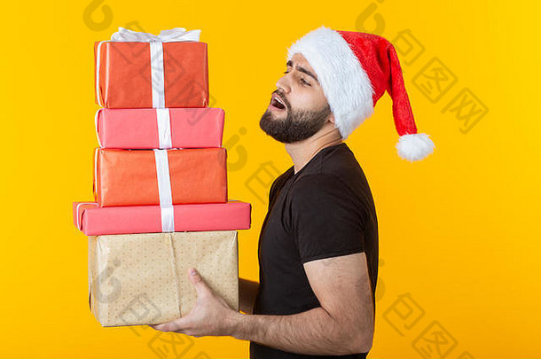 戴着圣诞老人帽子、留着胡子、<strong>心怀</strong>不满的年轻人拿着五个礼盒，在黄色背景上摆姿势。礼物和问候的概念