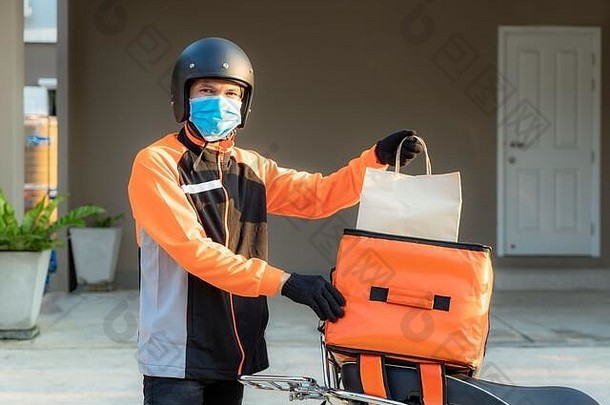 送货员亚洲男子戴着橙色制服的防护面具，准备将送货食品袋送到顾客家门口，并将箱子装在滑板车上，e