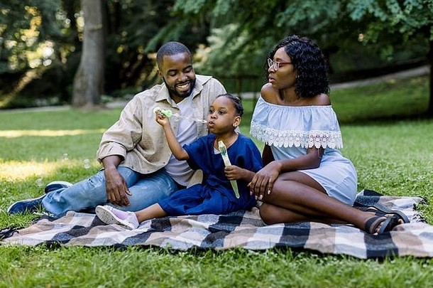 家庭、亲子关系和人的概念。快乐微笑的非洲家庭，爸爸妈妈和可爱的小女孩一起在公园里，在一个阳光明媚的日子里，吹着