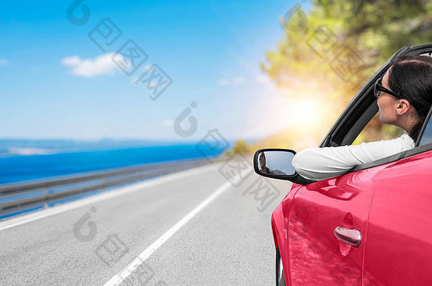 在一个阳光明媚的日子里，一名年轻女子坐在通往海边的路上的汽车里，背景是美丽的树木。