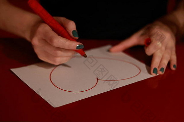 女孩用红色记号笔在白纸上画一颗心。手上有绿色指甲和记号笔。桌子上的白纸和图纸。
