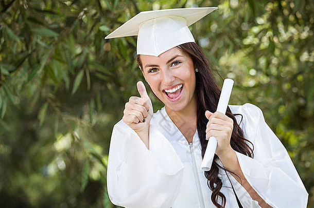 漂亮的混血女孩在外面戴着帽子，穿着长袍，手里拿着毕业证书庆祝毕业。