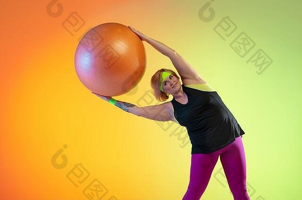 年轻的白人加大体型女模特在霓虹灯下的<strong>渐变橙色</strong>背景上训练。用健身球做运动。体育观念、健康生活方式、身体积极、平等。