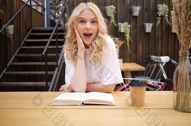 在城市咖啡馆的院子里，一个迷人的金发女孩拿着书和咖啡在照相机里惊奇地看着
