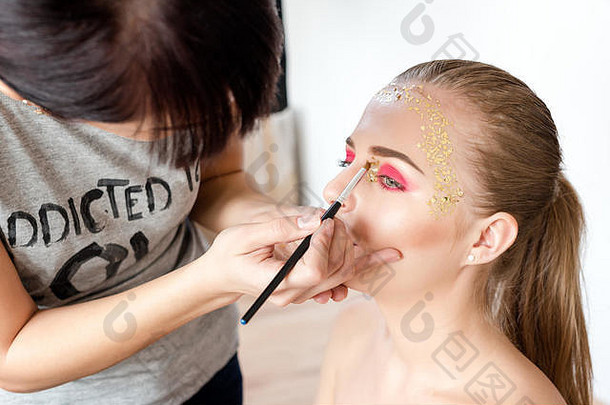 专业化妆师与美丽的金发女郎合作。后台场景：创意化妆大师应用眼影。漂亮的女人脸。化妆细节。皮肤完美的美女