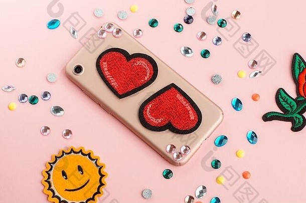 米色手机壳上的莱茵石和红色心形贴片。粉红色背景上的各种工艺材料。